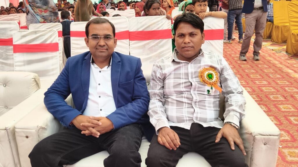 Principal Abhishek Srivastava seated on the stage sofa beside a guest wearing a ceremonial rosette, with the audience behind.