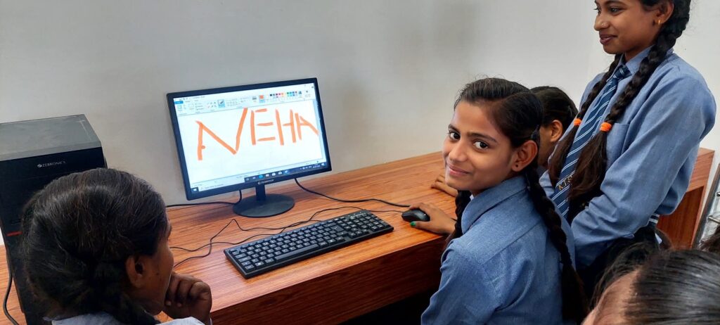 Students of the college seated at a desktop computer in the computer lab, with classmates watching over their shoulders.