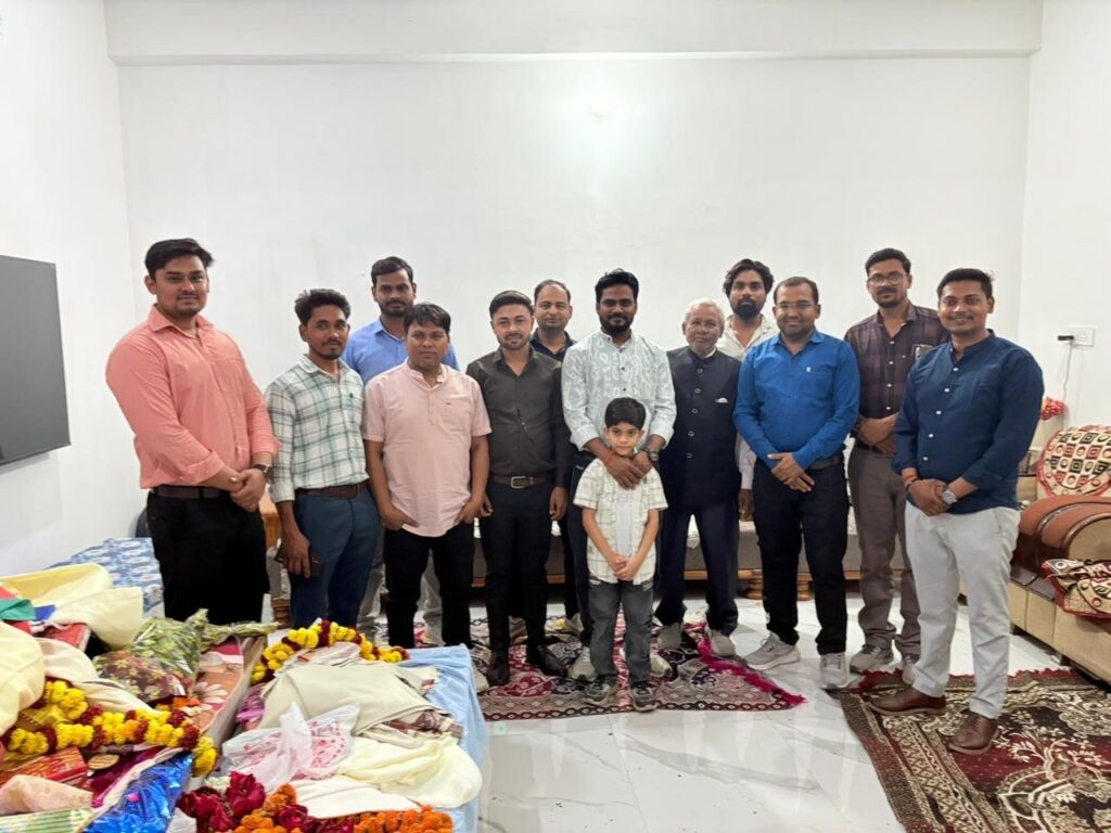 Group photograph of men at a gathering with garlands and wrapped gifts arranged in the foreground
