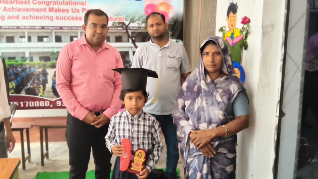 A primary-section student in a graduation cap holding a trophy, standing with his parents and college staff in front of a congratulations banner.