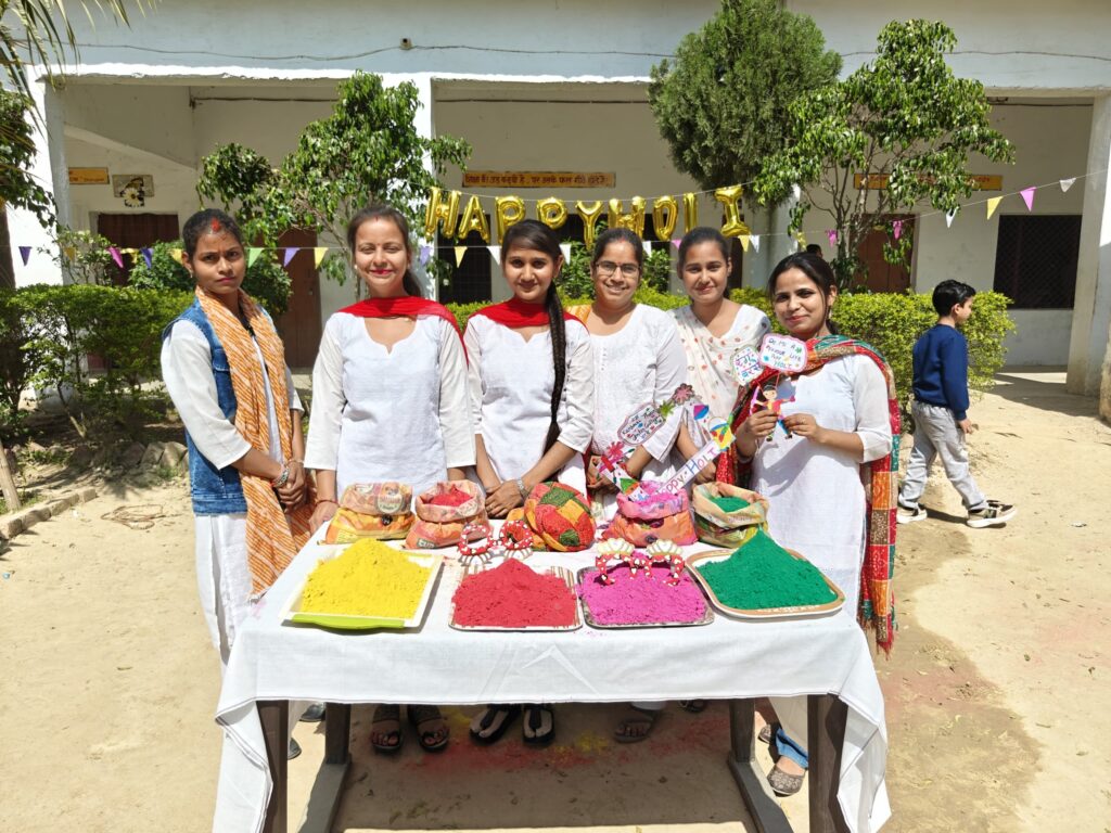 Women staff members of the college standing behind a table laid out with trays of yellow, red, pink and green Holi colours, under a Happy Holi balloon banner.