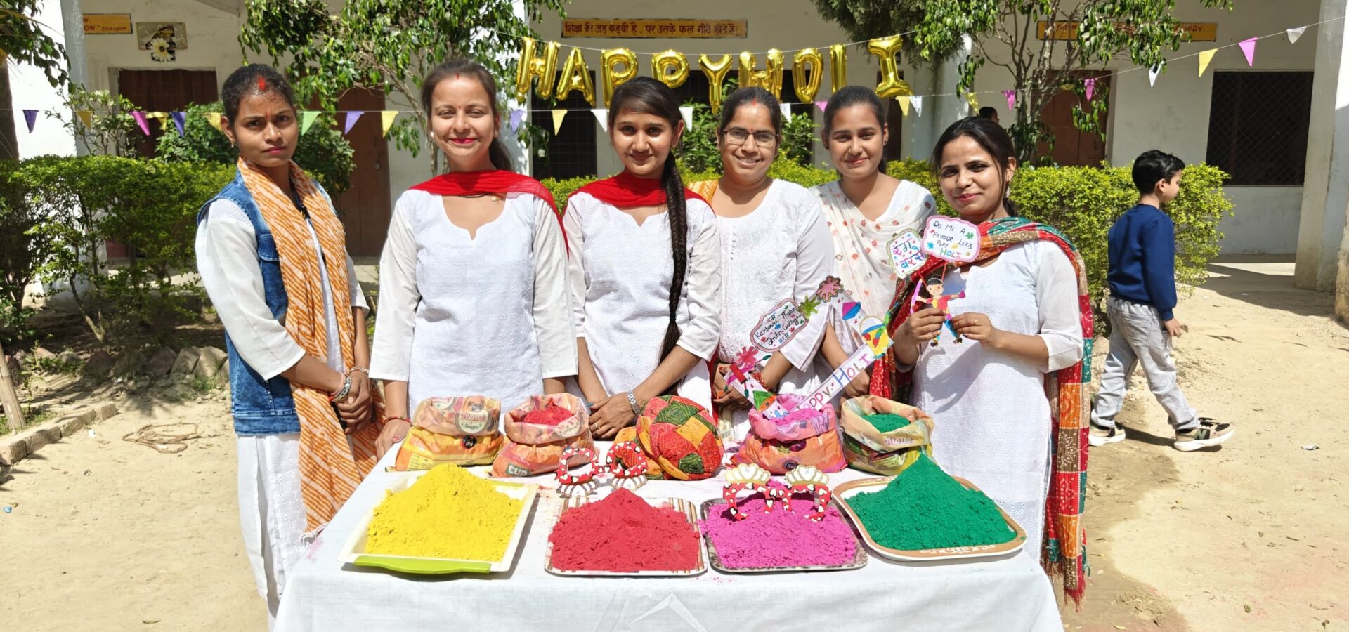 Women staff members of the college standing behind a table laid out with trays of yellow, red, pink and green Holi colours, under a Happy Holi balloon banner.