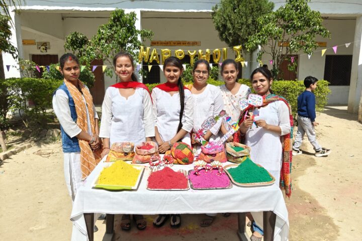 Women staff members of the college standing behind a table laid out with trays of yellow, red, pink and green Holi colours, under a Happy Holi balloon banner.