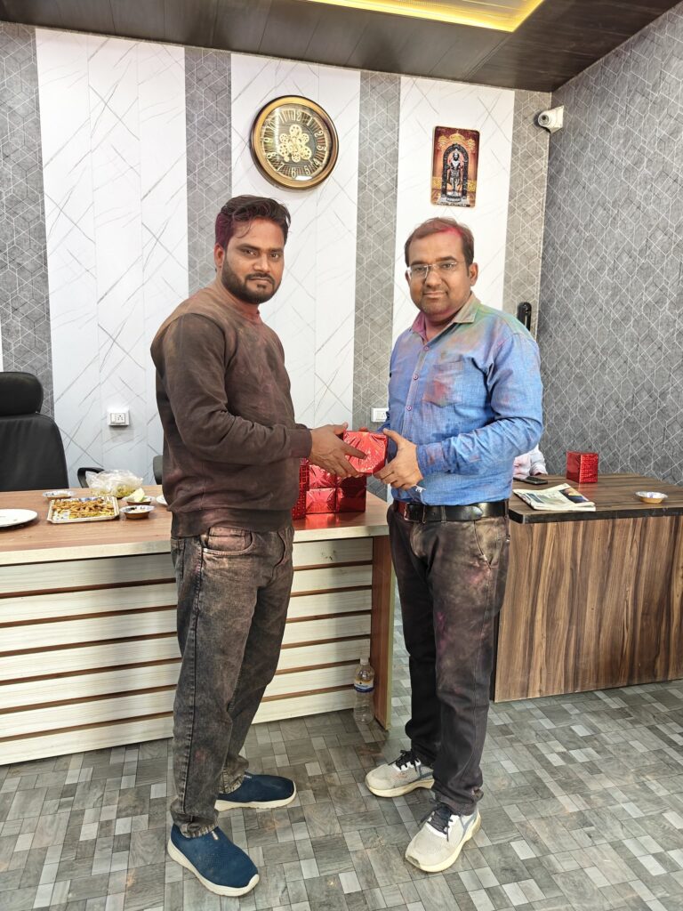Principal Abhishek Srivastava and Manager Hariom Sharma, their clothes marked with colour, exchanging a wrapped Holi gift in the college office.