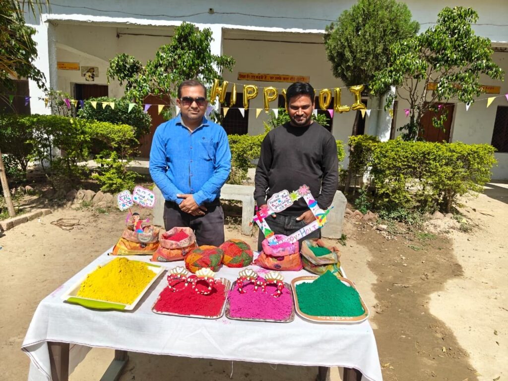 Two members of the college staff standing behind the Holi colour table in the courtyard, with Happy Holi balloons on the wall behind them.