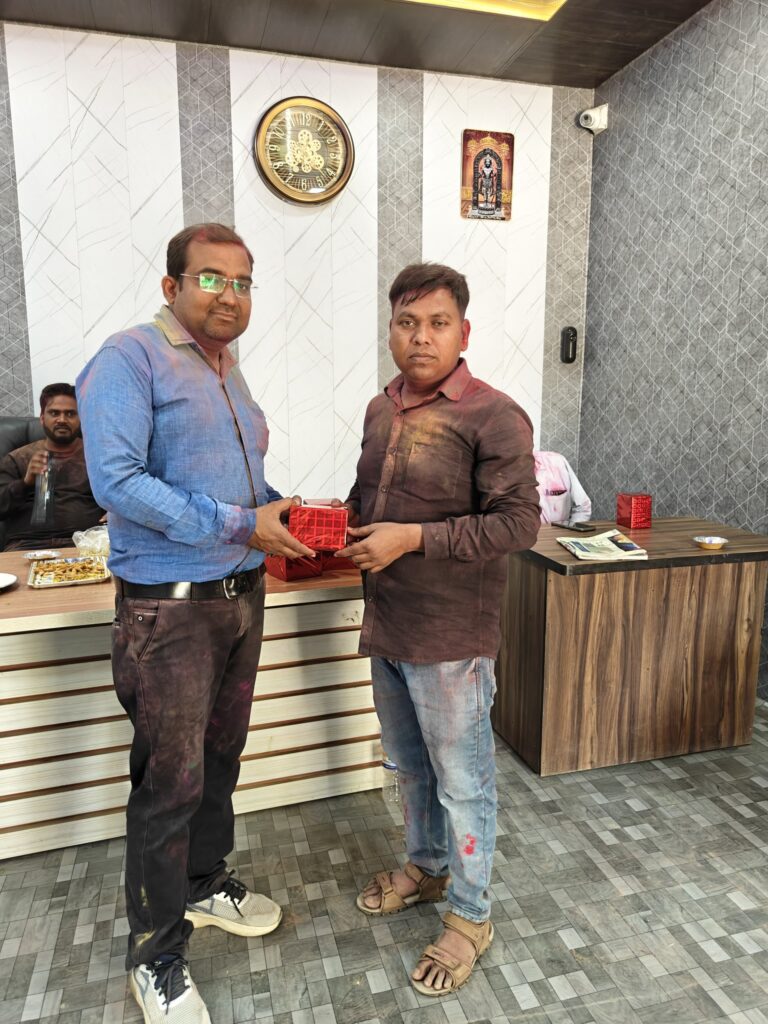 Principal Abhishek Srivastava presenting a wrapped Holi gift to Manager Hariom Sharma, both smeared with colour, in the college office.