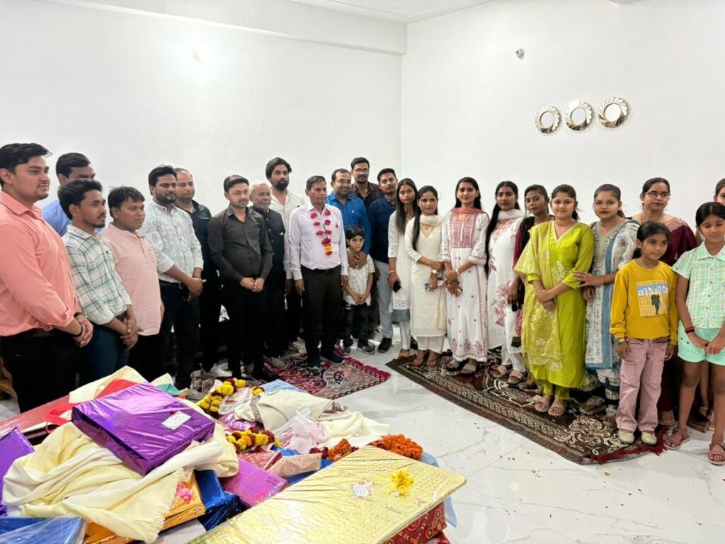 A large Holi Milan gathering of college staff and their families, with gifts and garlands laid out on the floor in front of them.