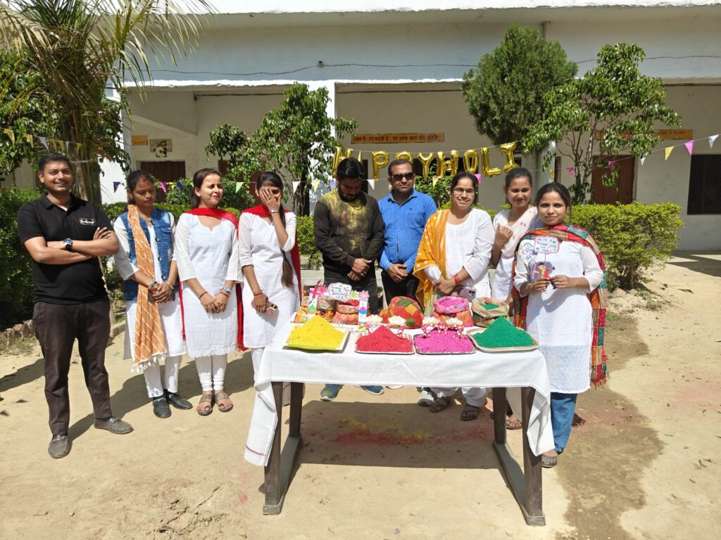 College staff standing behind the Holi colour table in the courtyard, with trays of coloured powder and Happy Holi balloons behind.
