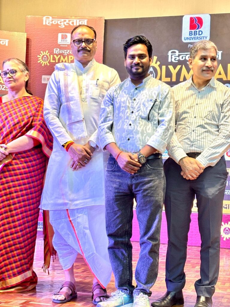 School staff standing with guests on stage in front of the Hindustan Olympiad 2025 backdrop
