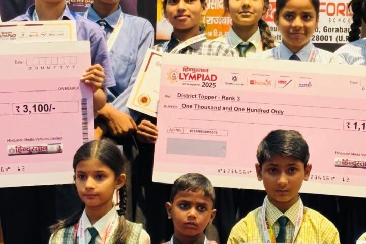 Students of Karpoori Thakur Inter College holding oversized Hindustan Olympiad 2025 prize cheques, including a District Topper Rank 3 award, along with merit certificates and medals.