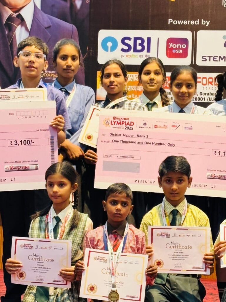 Students of Karpoori Thakur Inter College holding oversized Hindustan Olympiad 2025 prize cheques, including a District Topper Rank 3 award, along with merit certificates and medals.