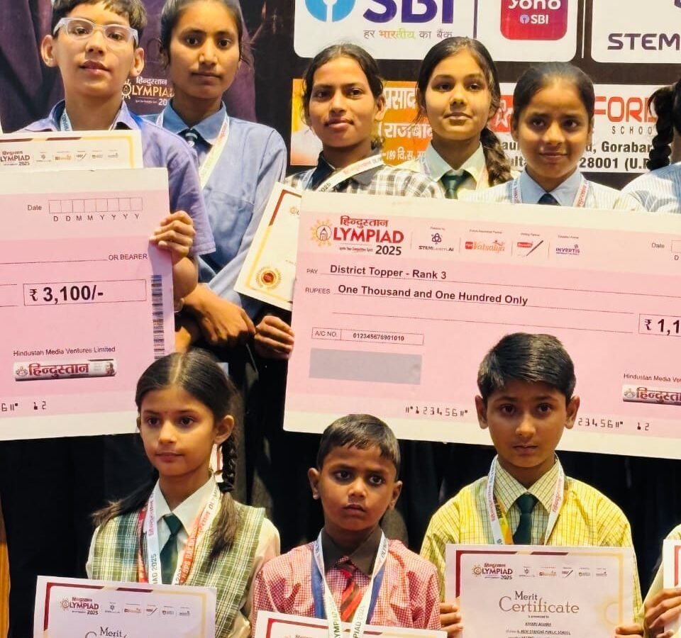 Students of Karpoori Thakur Inter College holding oversized Hindustan Olympiad 2025 prize cheques, including a District Topper Rank 3 award, along with merit certificates and medals.
