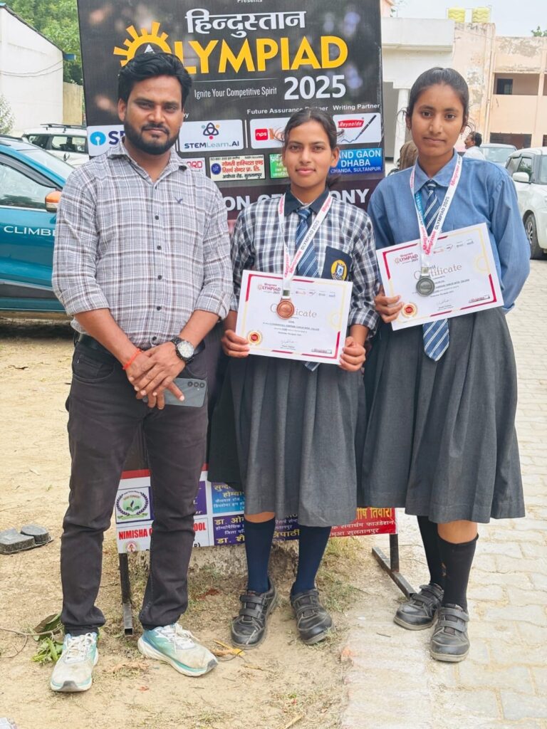 A teacher standing with two students holding Hindustan Olympiad 2025 merit certificates and medals in front of the felicitation ceremony backdrop.