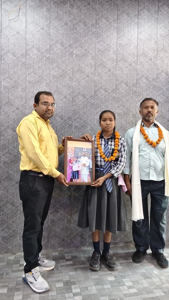 Principal Abhishek Srivastava holding a framed felicitation photograph beside a garlanded student and her garlanded father.