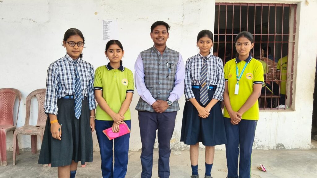 A teacher standing with four students, two in the blue check uniform and two in sports kit, outside a classroom.