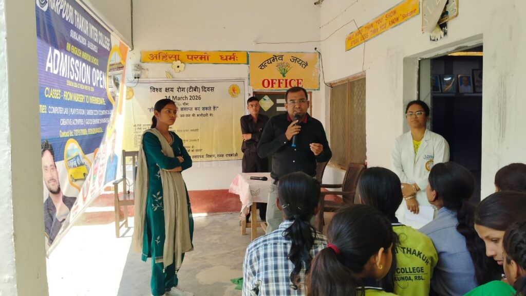 A speaker addressing students seated on the floor at the World TB Day awareness programme, with a doctor in a white coat standing alongside