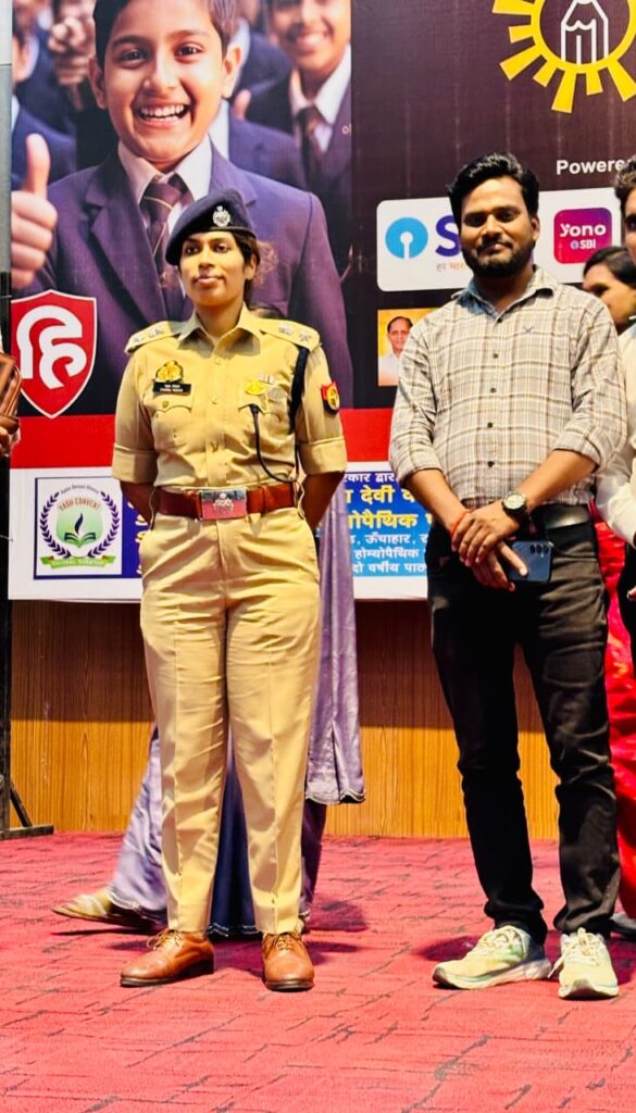 A member of the Karpoori Thakur Inter College staff standing on stage with a woman police officer in front of the Hindustan Olympiad 2025 backdrop