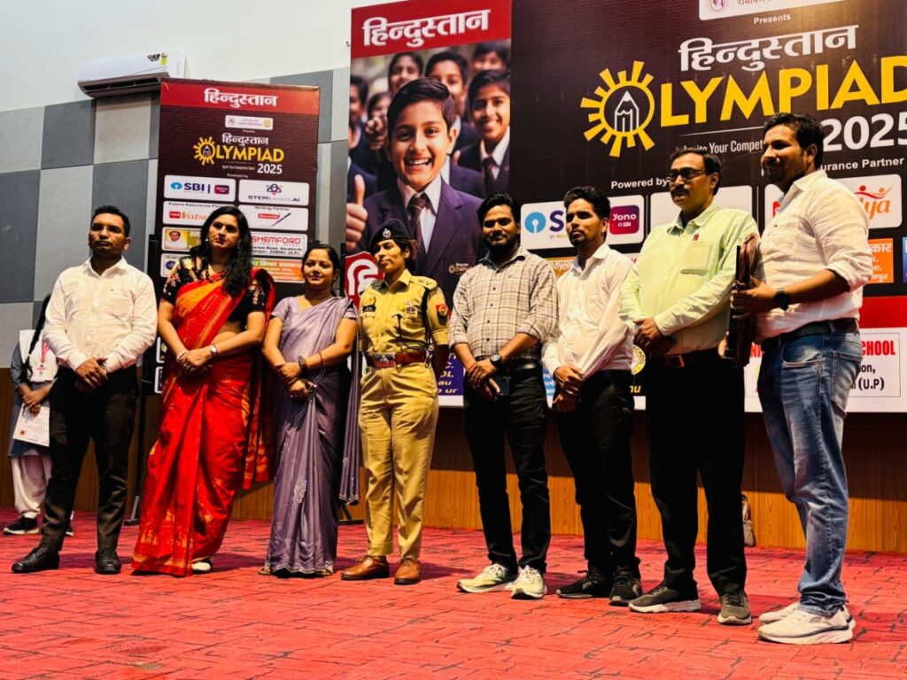 School staff and invited guests, including a police officer, standing together on stage in front of the Hindustan Olympiad 2025 backdrop