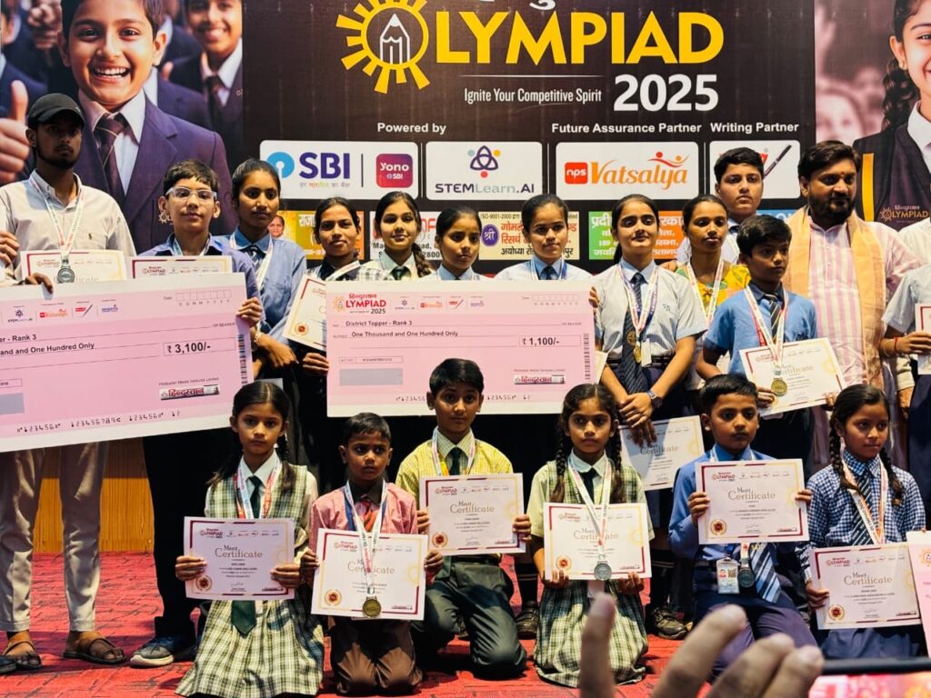 Hindustan Olympiad 2025 winners holding oversized District Topper prize cheques and merit certificates on stage at the award ceremony
