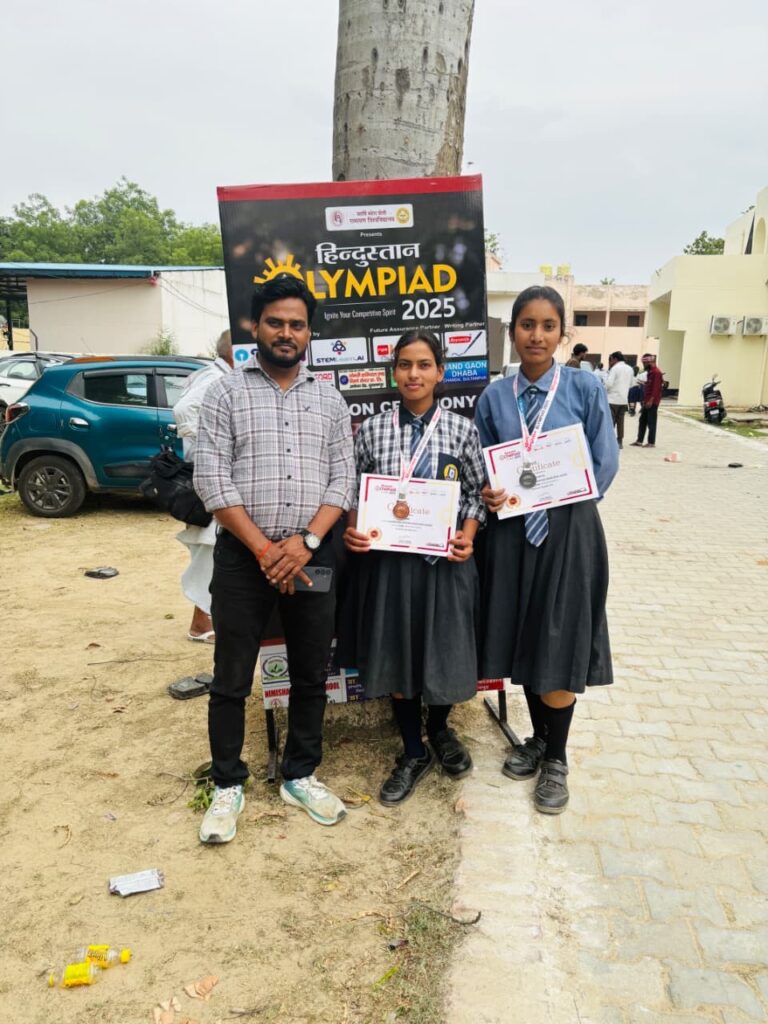 A teacher from Karpoori Thakur Inter College standing with two students holding Hindustan Olympiad 2025 certificates and medals beside the event standee outdoors